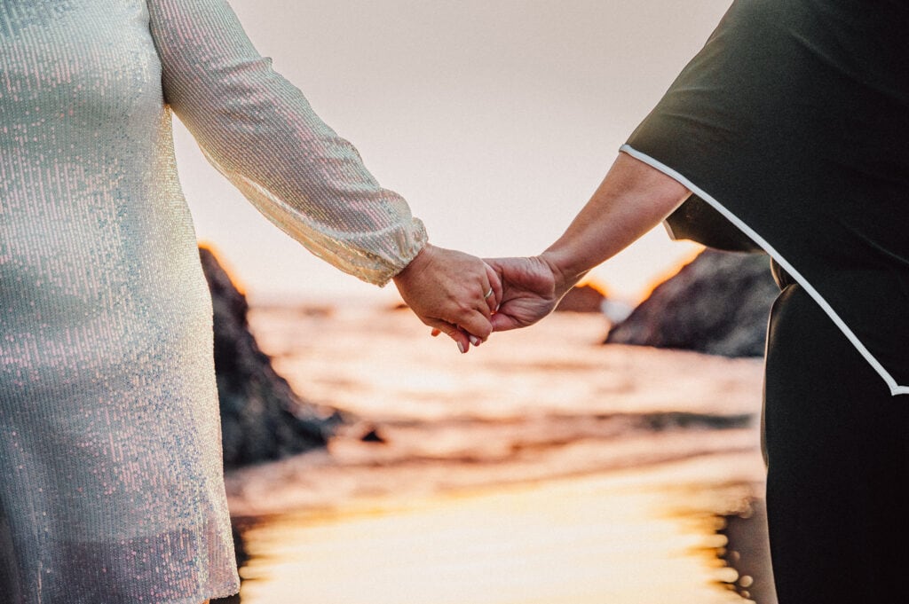 A close-up image of a newly wed couple holding hands with the sunset and beach in the background