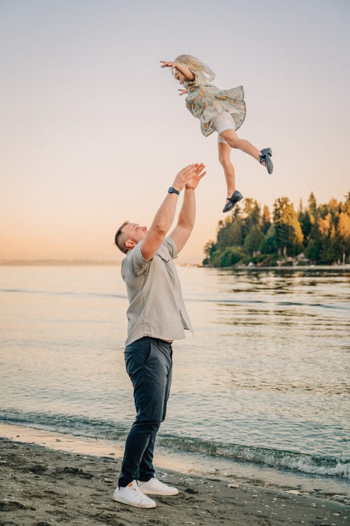 A father throws his young daughter in the air. The sun is setting over the water in Suquamish, WA
