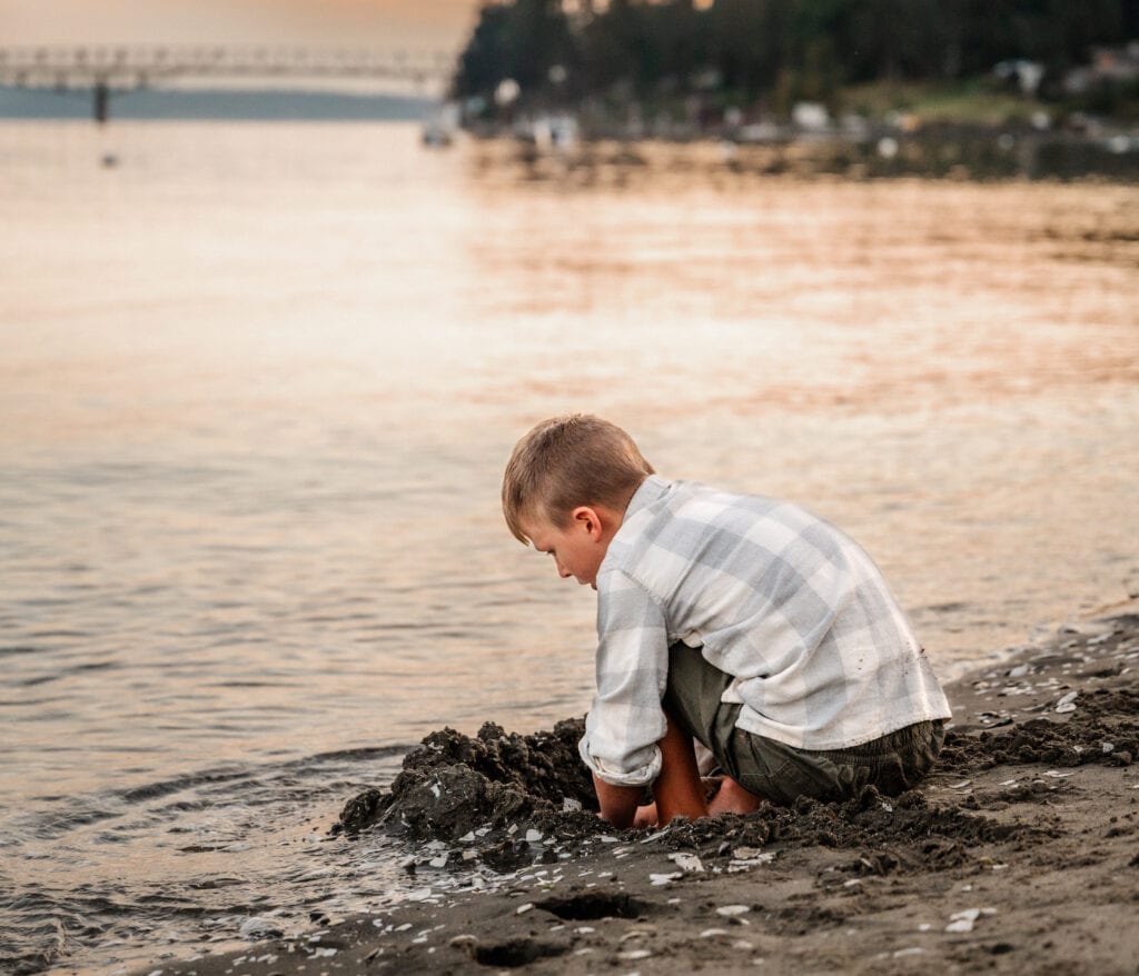 A boy digs in the sand at Old Man Beach. You can see the water and the Agate Pass bridge in the background