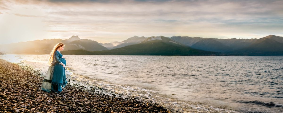A landscape portrait of a pregnant woman holding and looking at her belly. The Olympic Mountains and Agate Pass are in the background, lit up by the sunset. Taken at Scenic Beach State Park