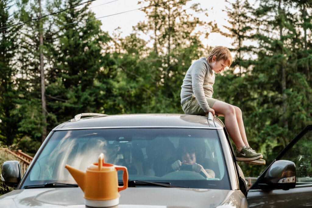 Candid image of an older child sitting on top of a car while their sibling smiles in the driver's seat. There is a yellow watering pitcher sitting on the car hood