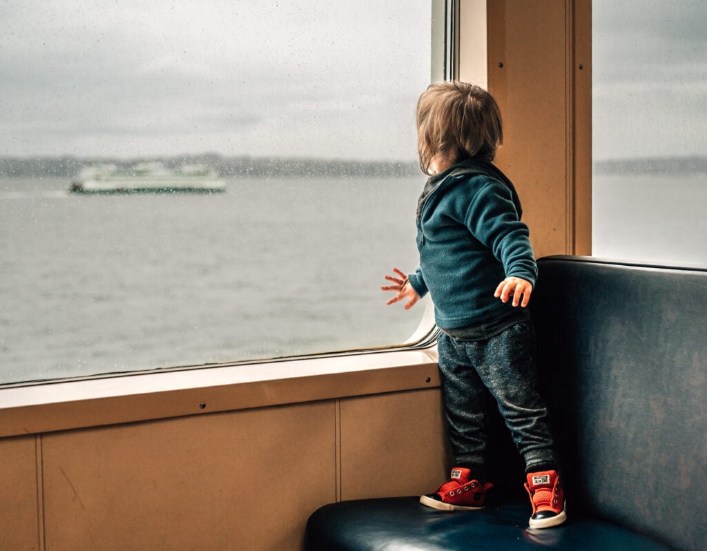 A child stands on a ferry seat and looks out the window at a passing ferry on the Edmonds-Kingston route