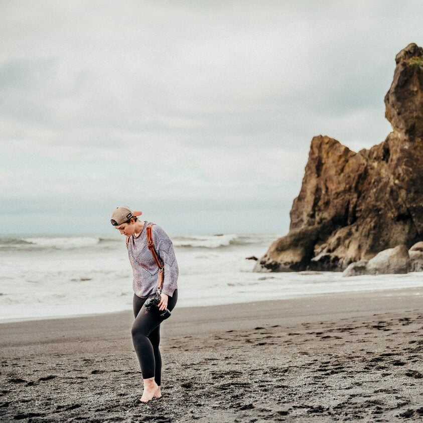 A person walking on a sandy beach near large rocks while looking down at the ground.