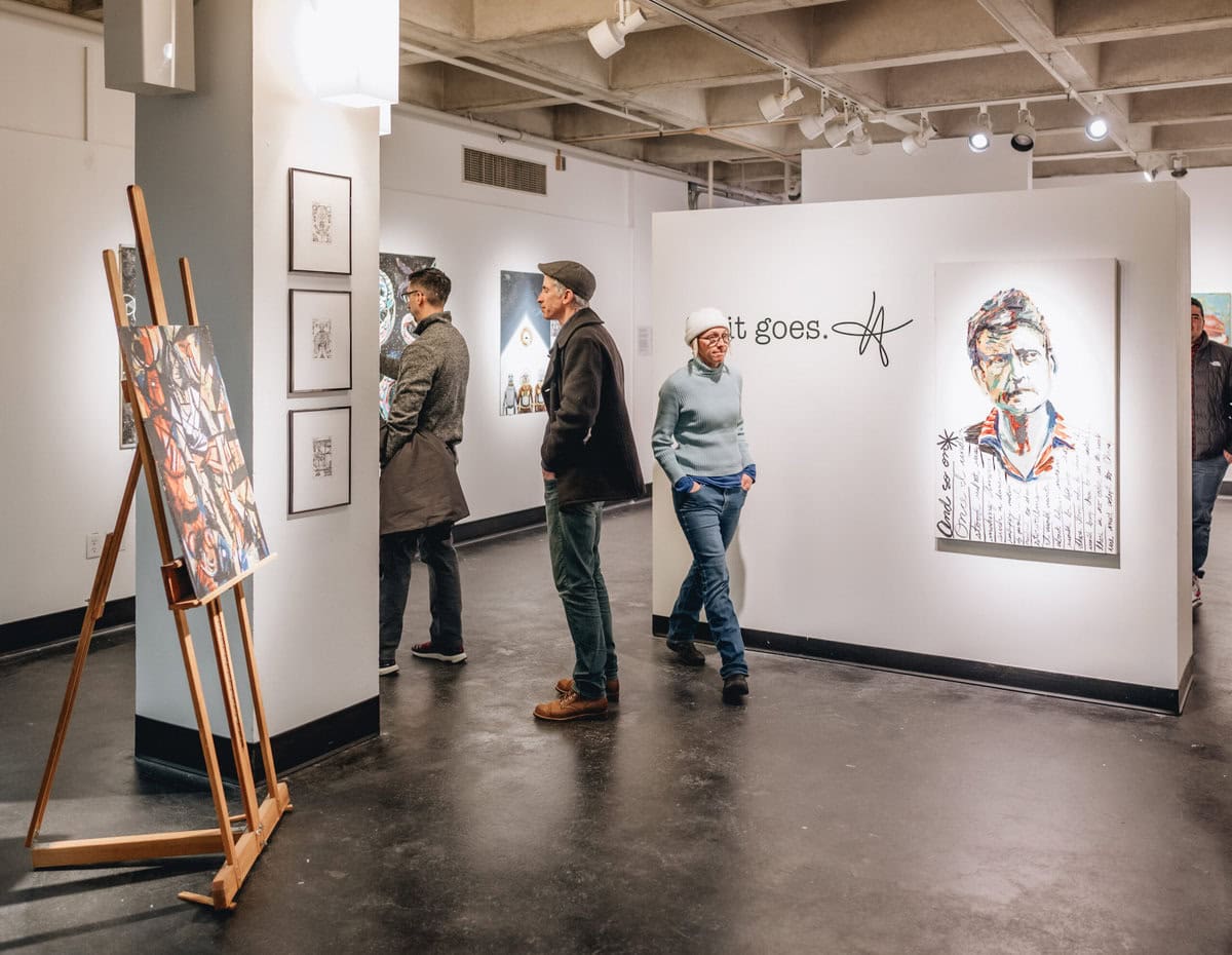 Visitors view artwork at a gallery exhibition featuring portraits and mixed media pieces.