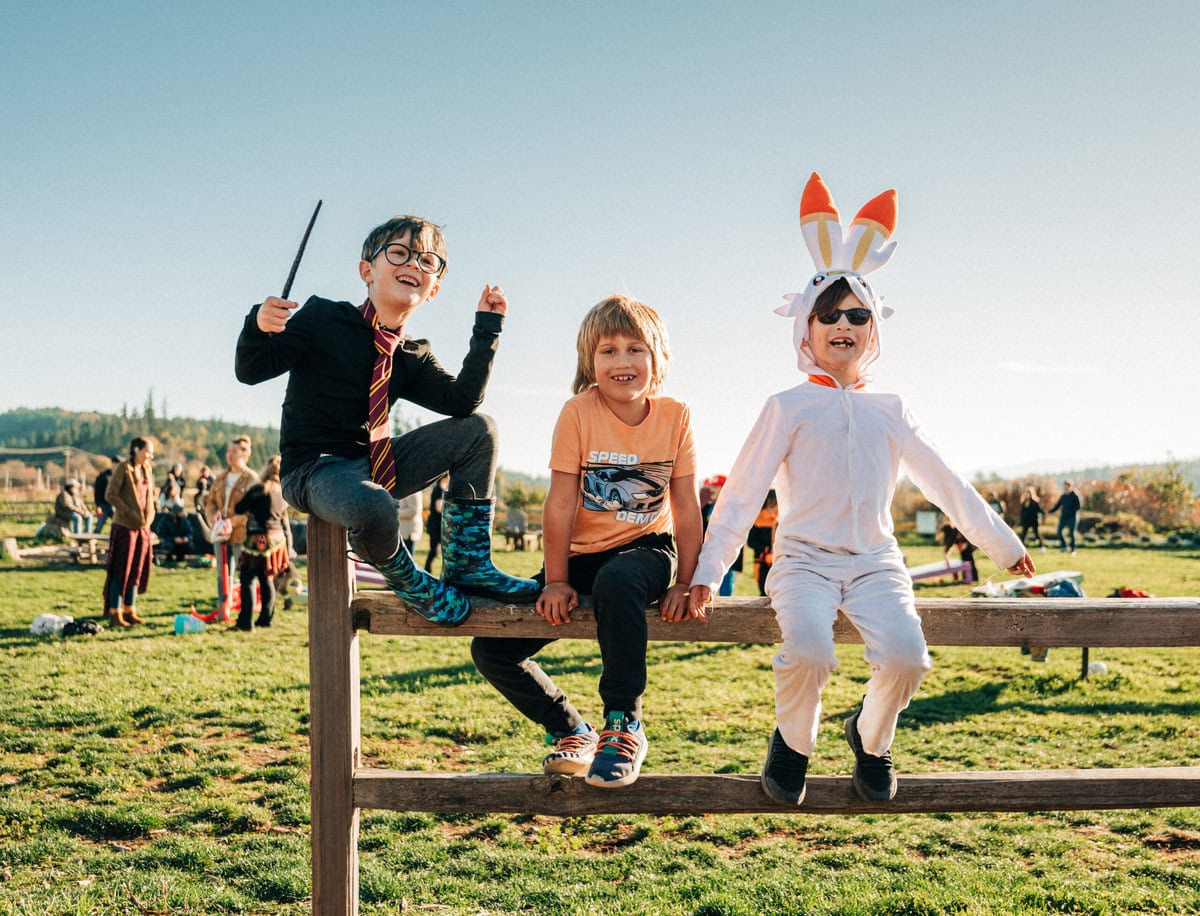 Three children in costumes playing on a wooden beam in a park during sunset, with one dressed as a wizard, another in casual wear, and the third in a bunny outfit.