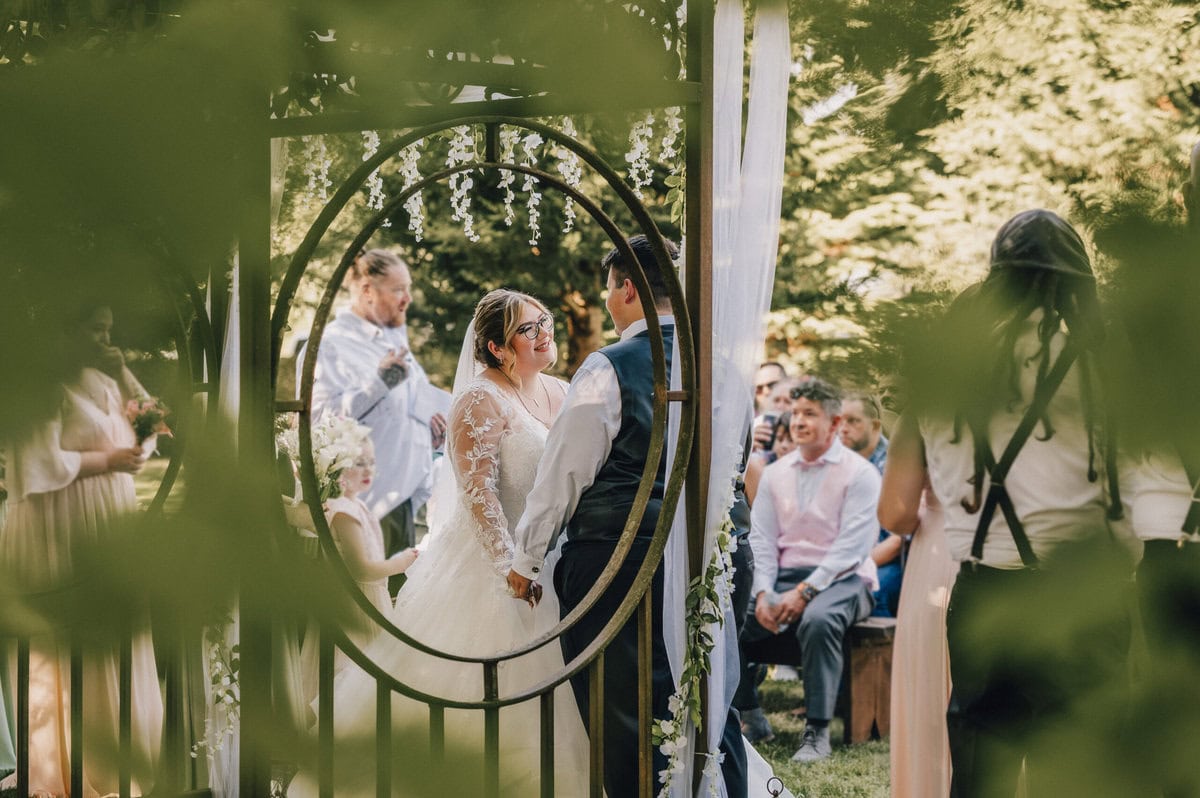 Bride and groom exchanging vows at an outdoor wedding ceremony, viewed through a circular gate surrounded by greenery.