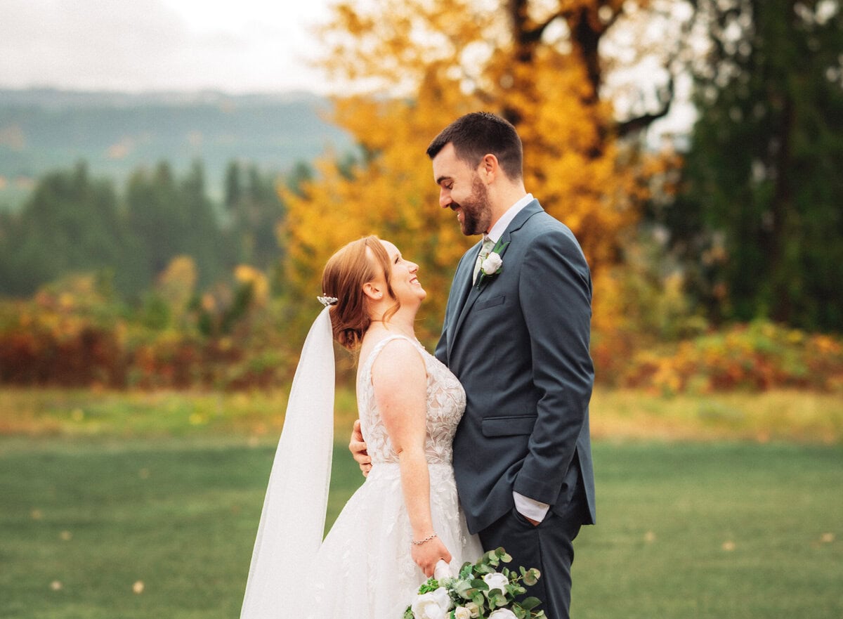 A bride and groom smiling at each other on a grassy field, surrounded by autumn trees with colorful leaves.