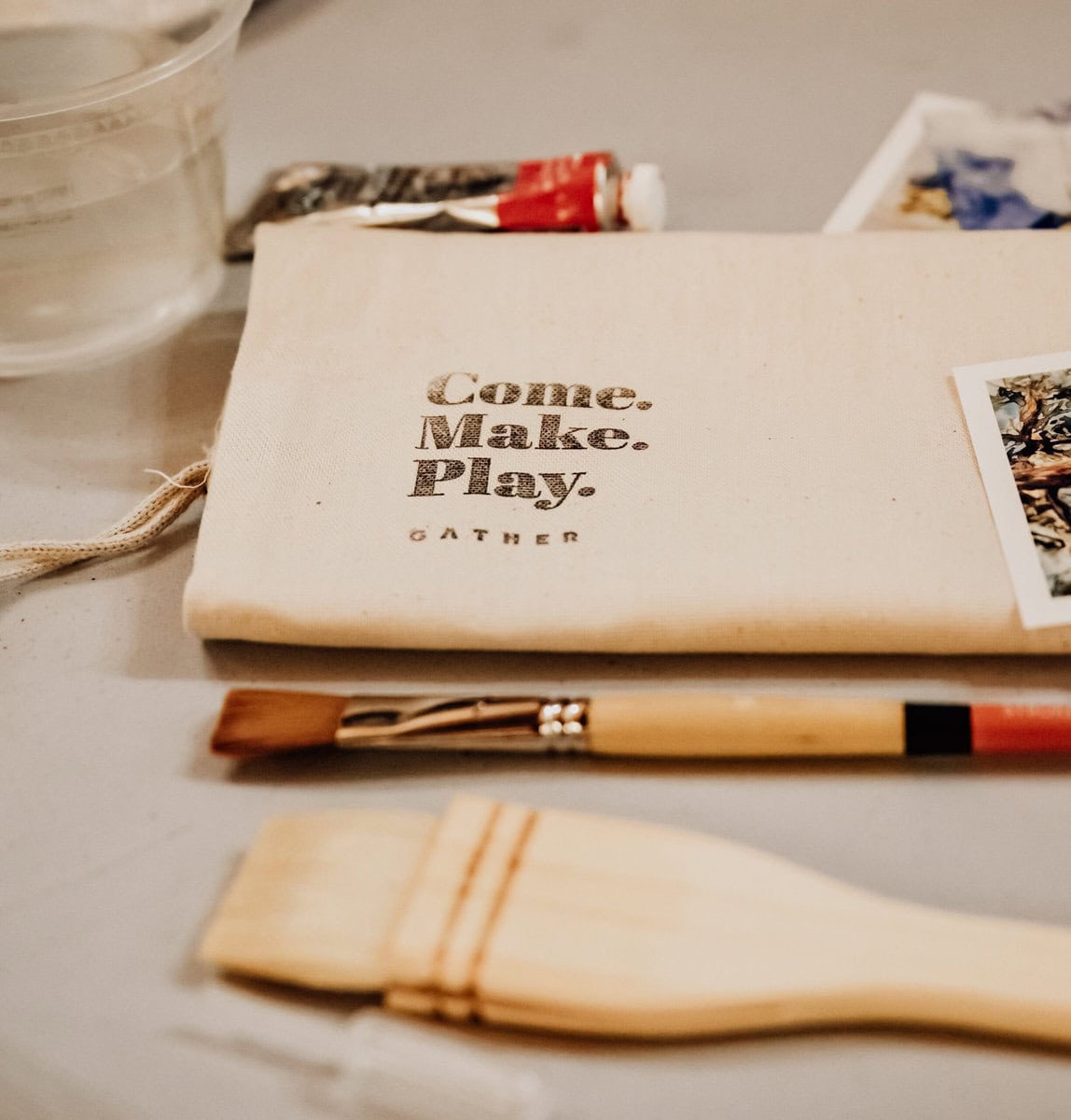 Art supplies including paint brushes and tubes on a table with a notebook that reads "come. make. play. gather.