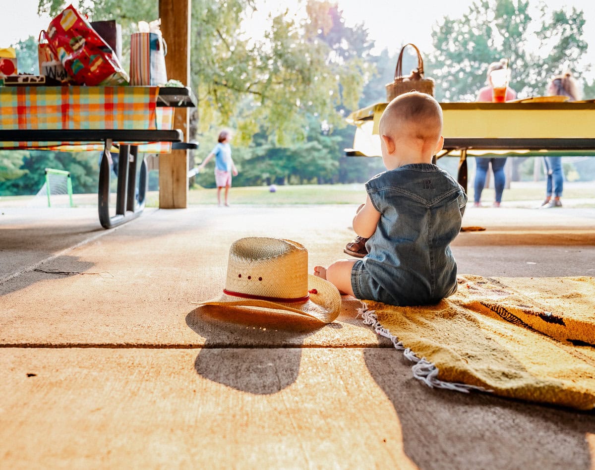 A baby in a denim outfit sits on a blanket at a sunny park, facing away from the camera toward a straw hat and a picnic table with people in the background.