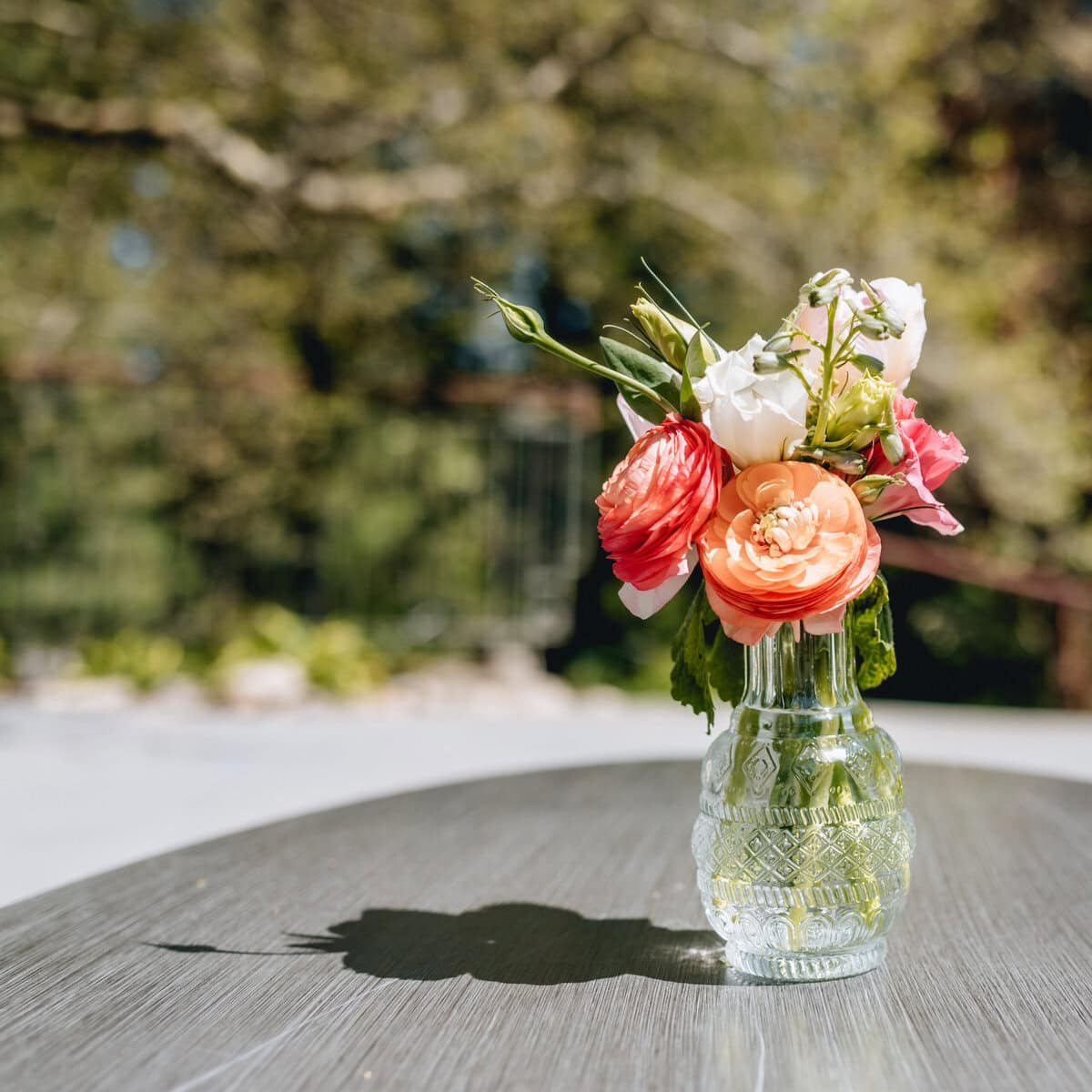 A bouquet of colorful roses in a glass vase on a wooden table outdoors, with natural sunlight creating a shadow of the vase on the table.